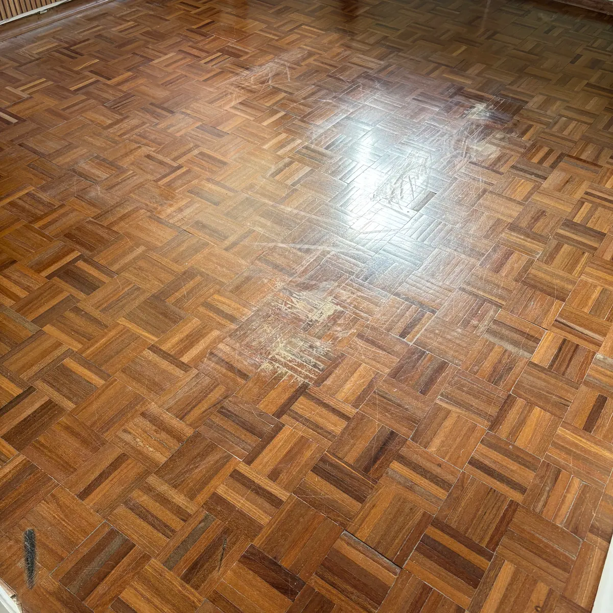 O mesmo piso de taco parquet antes da raspagem, com o verniz gasto, manchas e riscos